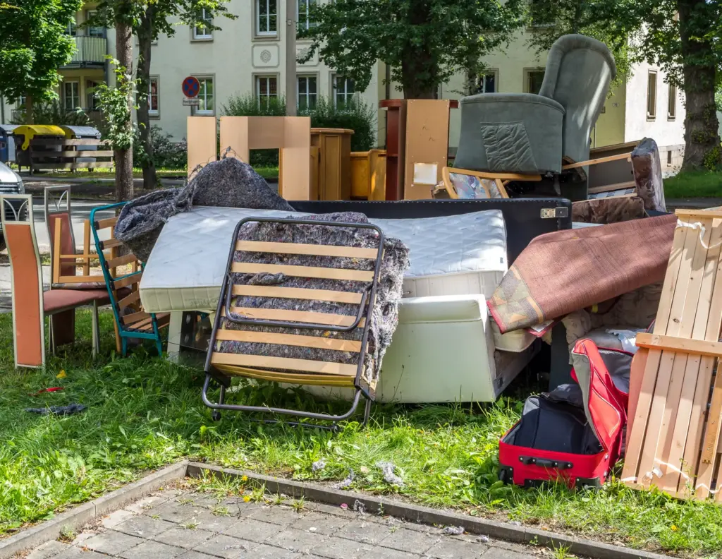 Matratzen, St&uuml;hle und Liegestuhl auf einem Sperrm&uuml;llhaufen unter B&auml;umen.