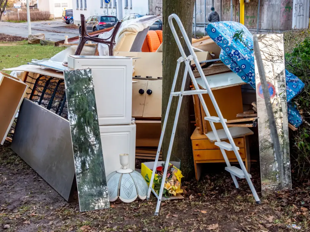 Ausrangierte M&ouml;bel, Leiter und Lampenschirm auf Sperrm&uuml;llhaufen vor B&auml;umen.