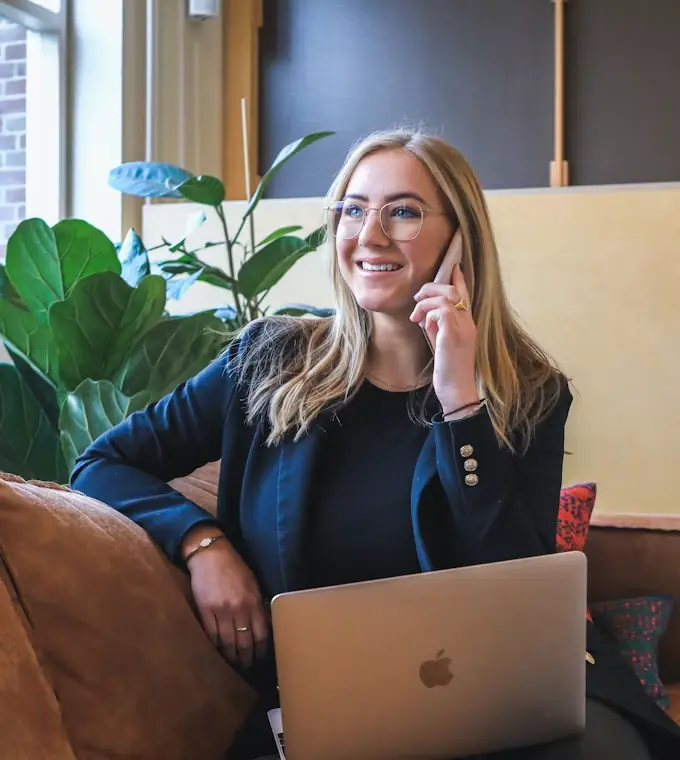 Woman with glasses smiling while talking on the phone, sitting on a couch with a laptop and a potted plant nearby.